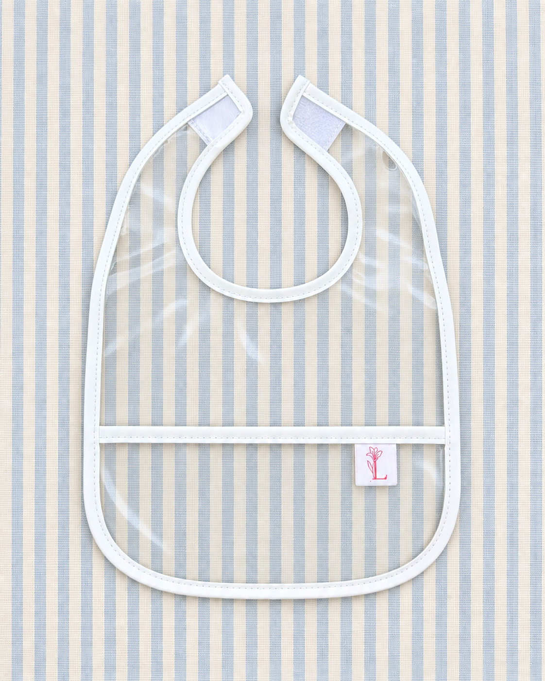 A clear plastic baby bib with white trim and straps, laid flat on a light blue and white vertically striped fabric background. A small white tag with a pink stylized 'L' logo is visible on the lower right of the bib.