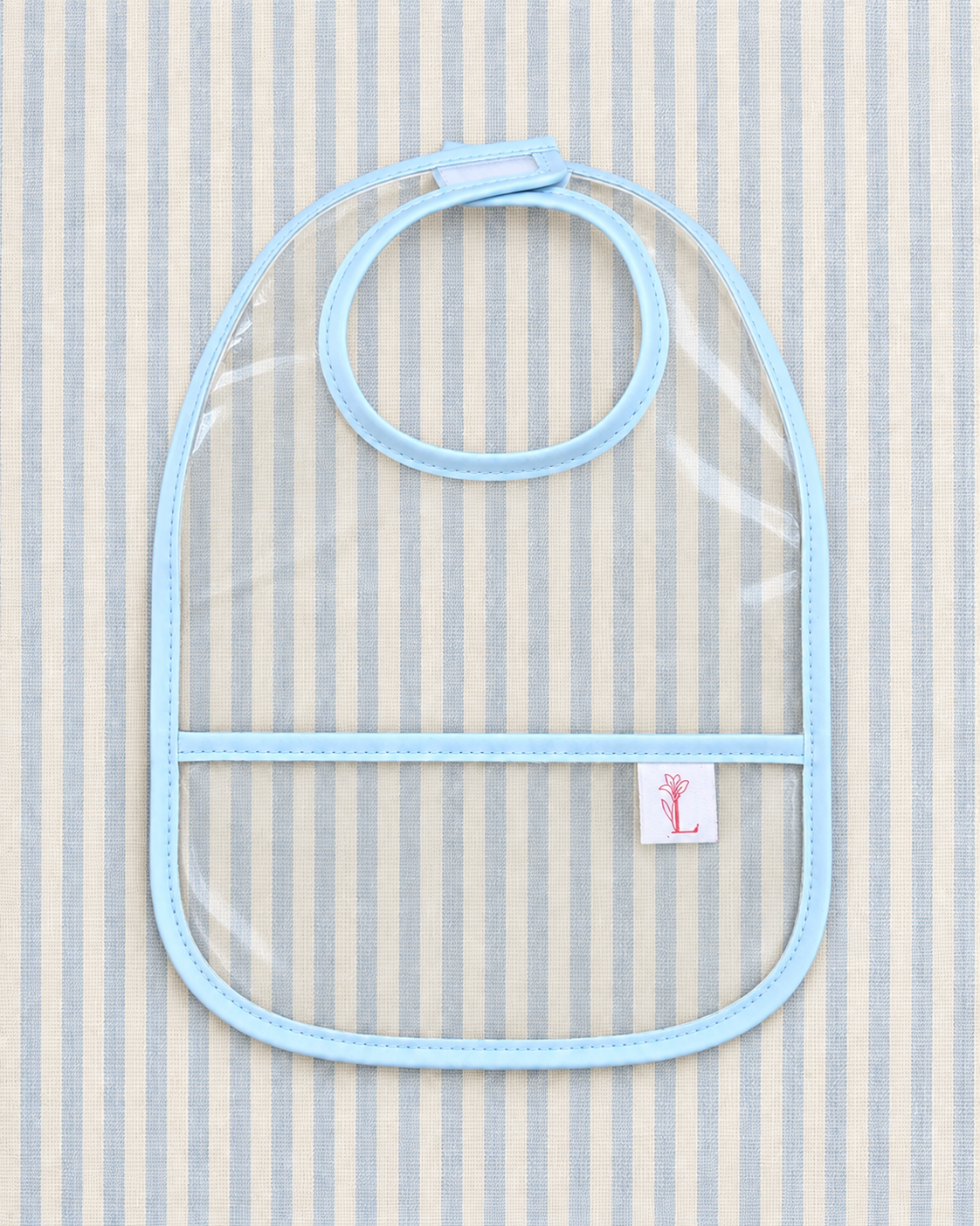 A clear plastic baby bib with light blue trim and a neck strap, laid flat on a light blue and white striped fabric background. A small white brand tag with a pink anchor logo is visible on the lower right.