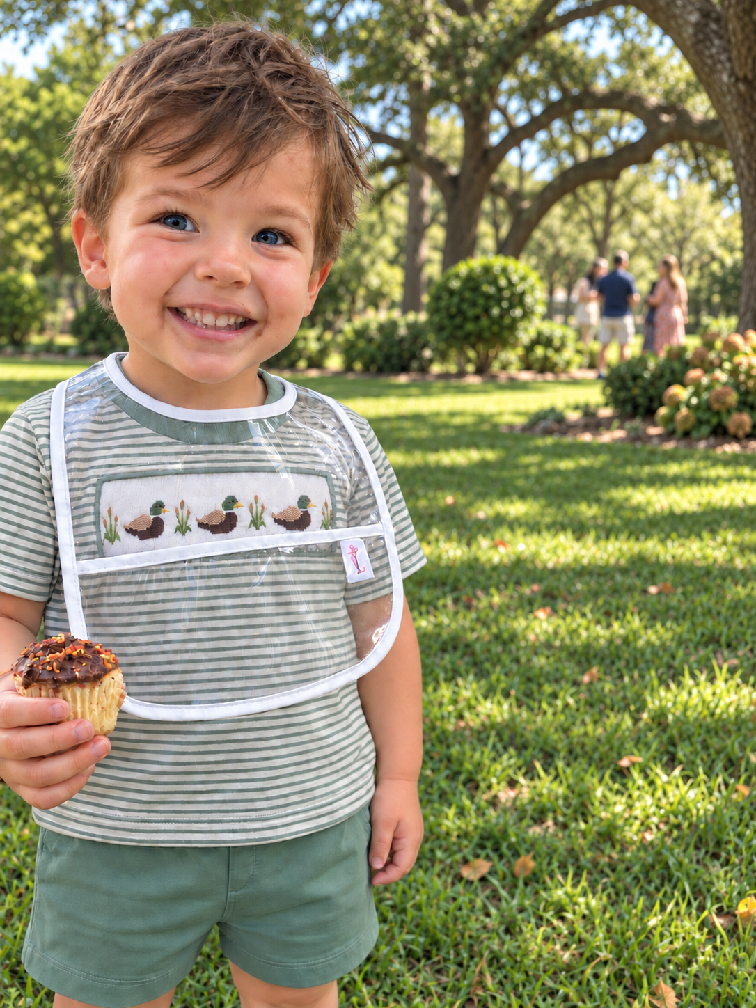 A smiling young boy standing outdoors on a grassy park lawn, wearing a clear invisibib bib with white trim and a duck-themed chest panel over a green striped shirt and green shorts, while holding a cupcake.